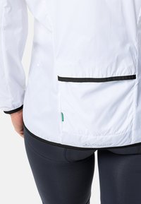 Leichte weiße Jacke mit schwarzen Akzenten, ausgestattet mit einer mit Reißverschluss versehenen Rückentasche und elastischen Bündchen, hergestellt aus glattem, wasserabweisendem Stoff.