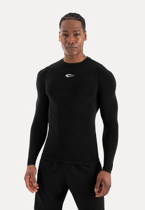 Uomo atletico con capelli intrecciati che indossa una maglia nera aderente a maniche lunghe e pantaloni neri, in piedi davanti a uno sfondo chiaro semplice.