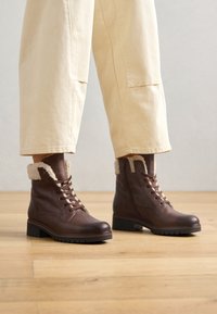 Botas de tobillo de cuero marrón con puños forrados de piel, suelas de goma y diseño de cordones. La textura es suave con un acabado mate.