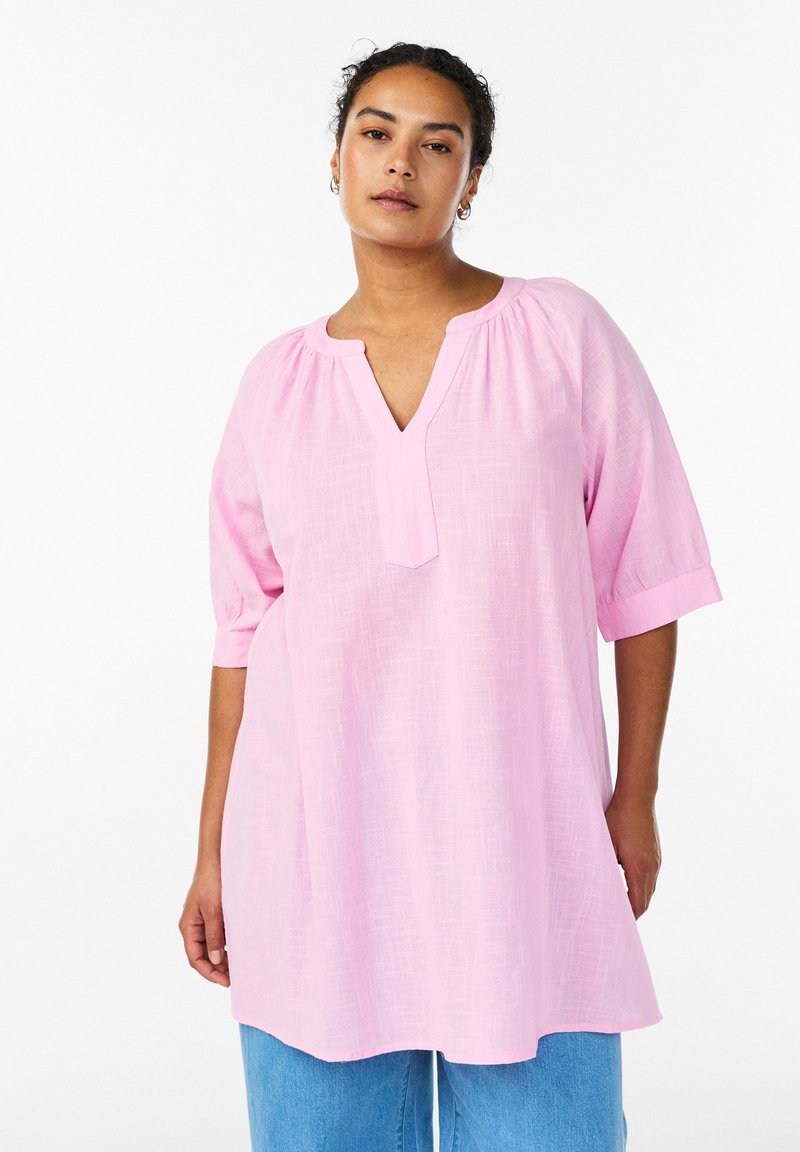 Hellrosa, locker sitzende Bluse mit V-Ausschnitt, Puffärmeln und strukturiertem Stoff. Der Saum reicht unter die Hüften und wird mit blauen Jeans kombiniert.