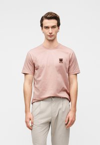 Ανοιχτό ροζ βαμβακερό μπλουζάκι με κοντά μανίκια και στρογγυλή λαιμόκοψη, με μικρό μαύρο λογότυπο στο στήθος. Συνδυασμένο με ανοιχτό γκρι παντελόνι.