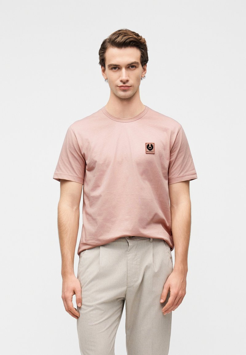 Ανοιχτό ροζ βαμβακερό μπλουζάκι με κοντά μανίκια και στρογγυλή λαιμόκοψη, με μικρό μαύρο λογότυπο στο στήθος. Συνδυασμένο με ανοιχτό γκρι παντελόνι.