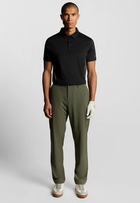 Sort poloshirt, olivengrønne bukser, hvite sko med gummisåle, iført en beige hanske. Enkel design med en tettsittende silhuett.