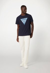 Marine T-shirt met een driehoekig blauw grafisch logo; gemaakt van zachte katoenen stof. Gecombineerd met crème kleurige, recht gesneden broek en witte sneakers.