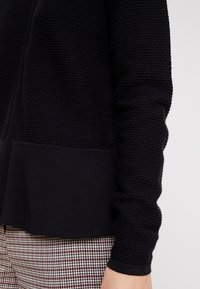 Schwarzer, strukturiert gestrickter Pullover mit glattem unterem Bund, ausgestattet mit einem durchgehenden Reißverschluss und gerippten Bündchen. Kombiniert mit karierten Hosen.