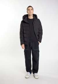 Sort pufferjakke med hætte, kviltet design, båret over en sort skjorte. Sammen med sorte cargo-bukser med hvid søm og sneakers.
