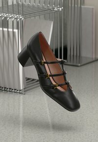 Black leather heeled shoe with three decorative straps and gold buckles. Square toe and block heel. Set against a minimalist background.