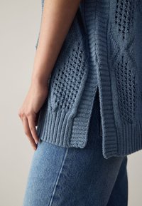 Pulover albastru deschis tricotat, cu modele texturate și fante laterale, asortat cu blugi din denim albastru. Prim-plan cu detalii ale mânecii și tivului.