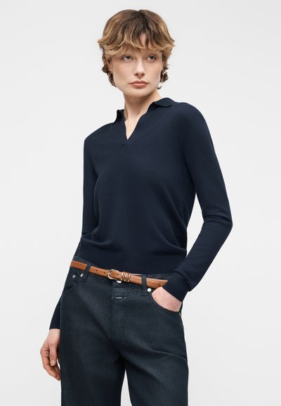 Jeune personne aux cheveux courts portant un haut à manches longues bleu marine foncé rentré dans un jean foncé avec une ceinture en cuir marron, main dans la poche.