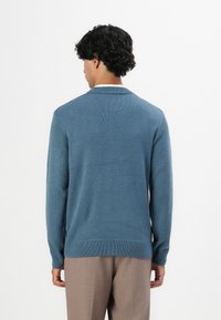 Man met krullend zwart haar, gekleed in een blauwe gebreide trui en beige broek, staande met zijn rug naar de camera tegen een witte achtergrond.