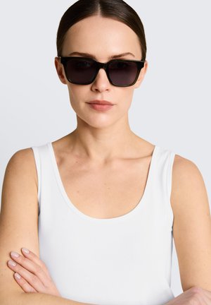 Femme à la peau claire et aux cheveux bruns, portant un débardeur blanc et de grandes lunettes de soleil noires, bras croisés, sur un fond clair uni.