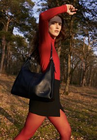 Žena v červenom top s dlhým rukávom a legínach, čiernej sukni, nesúca čiernu koženú kabelku cez rameno, kráča slnkom presvetleným lesom.