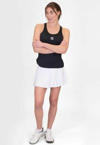 Top deportivo negro sin mangas con un logo, combinado con una falda blanca con pliegues. Llevado con zapatillas blancas con detalles en gris.