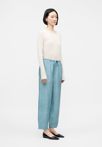 Γυναίκα με κοντά μαύρα μαλλιά, φορώντας λευκό μακρυμάνικο τοπ, ανοιχτόχρωμο μπλε παντελόνι με κορδόνια και μαύρα μυτερά παπούτσια, στέκεται μπροστά σε απλό φόντο.