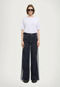 Dark blue wide-leg jeans with gray side panels, paired with a white t-shirt. Model wears black sunglasses and chunky black shoes.