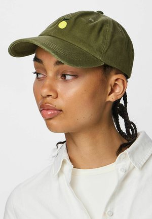 Ung person med flettet hår iført en olivengrøn kasket med en lille gul broderet frugt, hvid skjorte, kigger til siden.