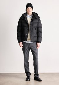 Μαύρο μπουφάν με υφασμάτινη υφή, συνδυασμένο με κρεμ πλεκτό πουκάμισο και μπλε ριγωτό πουκάμισο, γκρι παντελόνι και μαύρα παπούτσια.