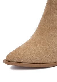 Beżowe zamszowe botki za kostkę z ostro zakończonym noskiem i płaską, jasną podeszwą. Gładka tekstura bez widocznych metalowych elementów czy wzorów.