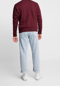 Burgundy sweatshirt med långa ärmar, rundhals. Ljust blå jeans med uppvikta ärmar. Vita sneakers. Motsatt vy som visar bakdetaljer.
