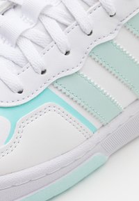 Biały sneaker z miętowozielonymi akcentami, teksturowaną skórą i warstwowymi paskami. Posiada białe sznurowadła oraz gumową podeszwę z teksturowaną powierzchnią.