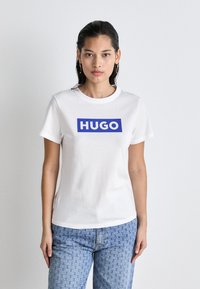 Bílý bavlněný tričko s modrým obdélníkovým logem "HUGO". Krátké rukávy a kulatý výstřih. Spárováno s kostkovanými modrými džíny.