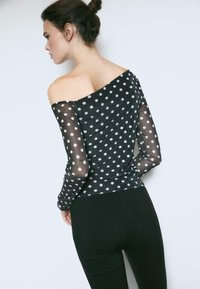 Schwarzes Off-Shoulder-Oberteil mit durchsichtigen Pünktchenärmeln; figurbetonte Silhouette; kombiniert mit schwarzen Leggings; glatte Textur.