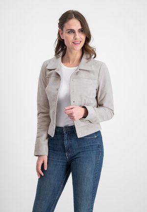 Frau trägt eine hellgraue kurze Jacke über einem weißen Hemd und blauen Jeans, lächelt und schaut leicht nach links vor einem weißen Hintergrund.