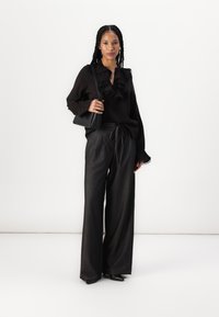 Blusa nera con dettagli a volant, abbinata a pantaloni ampi scuri. La modella tiene una piccola borsa nera. Texture lisce e vestibilità su misura.
