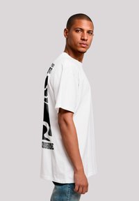 Weißes T-Shirt mit kurzen Ärmeln, das ein großes schwarzes Grafikdesign auf dem Rücken zeigt. Runder Ausschnitt und lässige Passform. Kombiniert mit blauen Jeans.