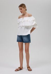 Off-the-shoulder witte blouse met gerimpelde lagen, gecombineerd met denim shorts. Model draagt bruine sandalen, heeft een gestructureerde stof.
