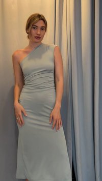Femme portant une robe asymétrique une épaule, sans manches, de couleur gris clair, debout devant des rideaux gris clair assortis.