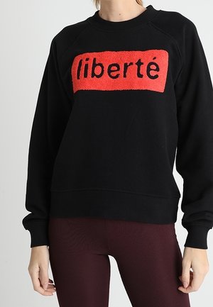 Sweat-shirt noir à manches longues avec un patch rectangulaire rouge affichant le mot « liberté », porté par une personne en pantalon côtelé bordeaux.