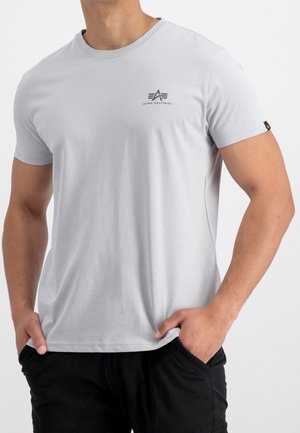 Homme portant un t-shirt gris clair Alpha Industries avec logo sur la poitrine, mains dans les poches, associé à un pantalon noir, sur fond uni.