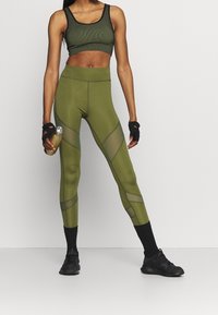 Reggiseno sportivo verde a costine con leggings alti coordinati. I leggings presentano pannelli trasparenti e un design geometrico. Scarpe da ginnastica nere e bottiglia d'acqua.