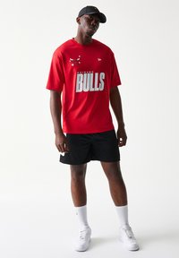 Κόκκινο μπλουζάκι με δίχτυ Chicago Bulls και λευκά γράμματα, συνδυασμένο με μαύρο σορτς και λευκά αθλητικά παπούτσια. Μαύρο καπέλο με λογότυπο.