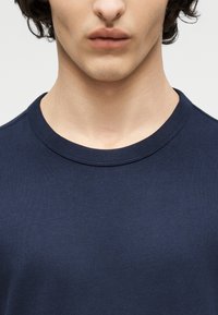 Νεαρός με ανοιχτόχρωμο δέρμα και σγουρά σκούρα μαλλιά, ντυμένος με μπλε σκούρο μπλουζάκι χωρίς γιακά, εμφανιζόμενος από τους ώμους μέχρι τα χείλη με φόντο λευκό.