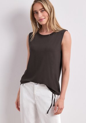 Femme souriante aux longs cheveux blonds portant un haut noir sans manches avec un nœud sur le côté et un pantalon blanc sur un fond clair uni.