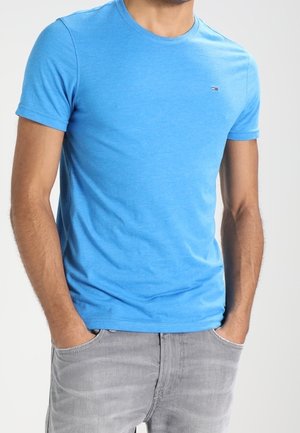 Jasnoniebieska bawełniana koszulka z okrągłym dekoltem i krótkimi rękawami, z małym logo na piersi. Noszona z szarymi dżinsami.