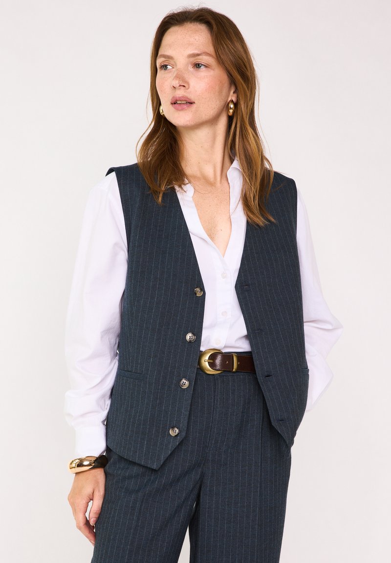 Gilet grigio a righe sottili con taglio sartoriale, bottoni argentati, indossato sopra una camicia bianca con colletto e completato da una cintura marrone.