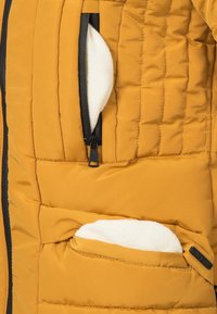 Pufferjack in mosterdgeel, met een doorgestikte textuur, twee ritszakken met witte fleece voering en zwarte ritsdetails.