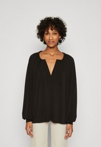 Svart langermet bluse med V-hals og rynkedetaljer ved halsåpningen. Myk tekstur og flytende design, paired med lyse bukser.