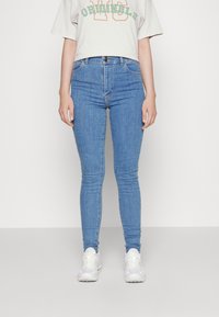 Jeans de tiro alto en azul claro con un diseño ajustado, que cuentan con un estilo clásico de cinco bolsillos y una cremallera. Combinados con una camiseta corta en gris claro.