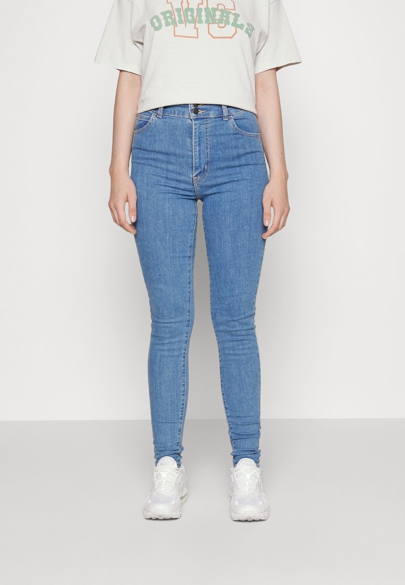 Jeans de tiro alto en azul claro con un diseño ajustado, que cuentan con un estilo clásico de cinco bolsillos y una cremallera. Combinados con una camiseta corta en gris claro.