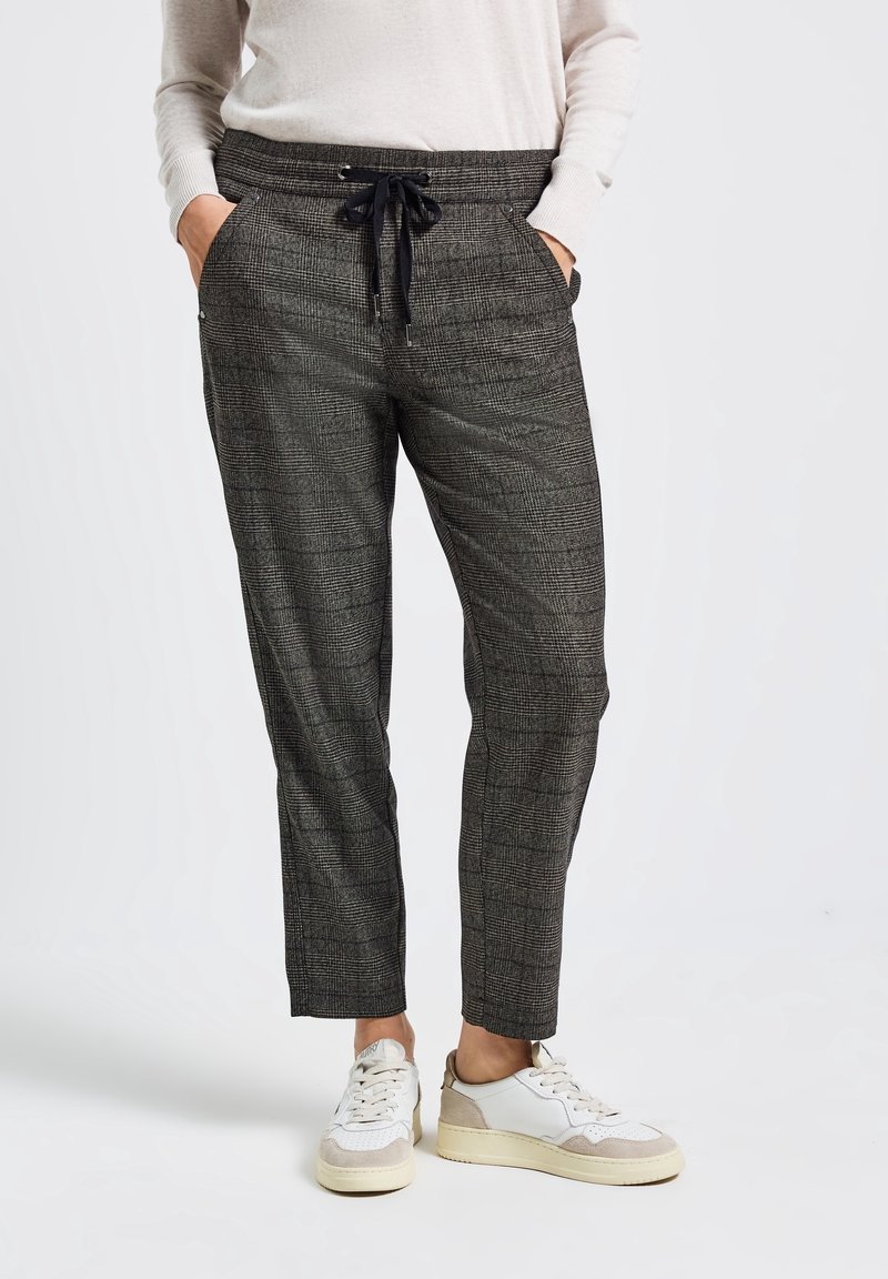 Personne portant un pantalon gris à carreaux avec cordon de serrage, les mains dans les poches et des baskets blanches, debout devant un fond uni.