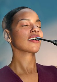Femme aux yeux fermés se brossant les dents de devant avec une brosse à dents électrique noire à tête de poils bleus.