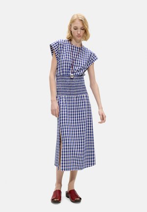 Młoda kobieta w niebiesko-białej sukience midi w kratę z bocznym rozporkiem, czerwonych drewniakach na odkryte palce i długim naszyjniku z srebrnym wisiorkiem.