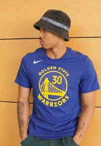 Camiseta de algodón azul con el logo amarillo de "Golden State Warriors" y un gráfico de un puente. Sombrero tipo cubo negro con una banda de patrón azul.