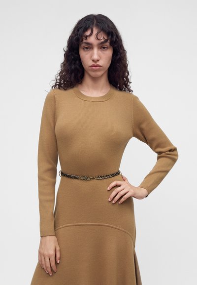 Un mannequin aux longs cheveux foncés et bouclés pose en robe en maille beige, cintrée par une ceinture chaîne, sur un fond uni gris clair.