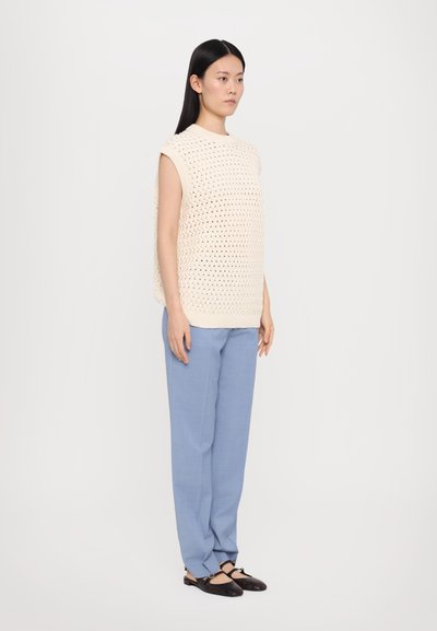 Svijetlosmeđi bezrukavnik s teksturiranim uzorkom, uparen sa svijetloplavim hlačama i tamnosmeđim cipelama, stoji naslonjen na bijelu pozadinu.