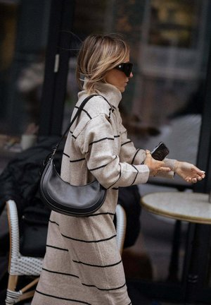 Beige trui jurk met zwarte strepen, hoge kraag en losse pasvorm; gecombineerd met een zwarte schoudertas. Accessoire: oversized zwarte zonnebril.
